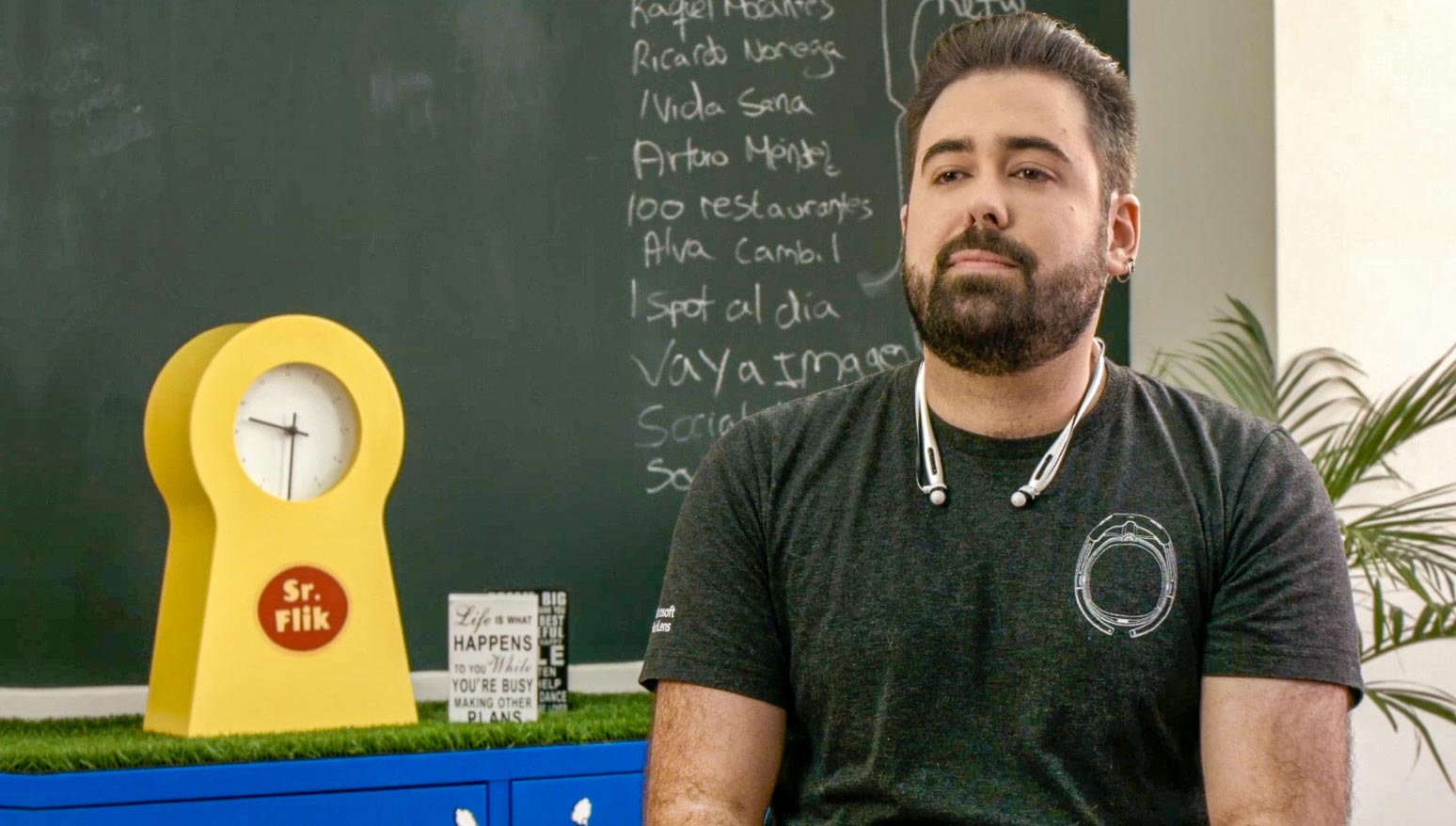 Un hombre con barba y auriculares alrededor de su cuello, de pie frente a un pizarrón.